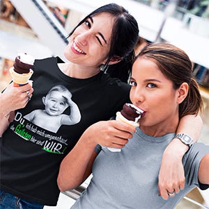 Zwei junge Frauen genießen Eis und tragen moderne T-Shirts mit kreativen Designs. Die Frau links trägt ein schwarzes T-Shirt mit einem lustigen Baby-Motiv und dem Spruch "Du bist nicht umsonst Chef, hier ist nur WIR!". Beide lächeln und wirken entspannt in einer lebhaften Umgebung, was den Alltagshumor und den Stil der T-Shirts unterstreicht.